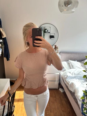 Ljusrosa croppad blus med volang - Söt ljusrosa croppad blus med volangkant och korta ärmar. Toppen har en lätt rynkad struktur och markerad midja som ger en feminin siluett. Perfekt för dig som gillar en romantisk och trendig stil.