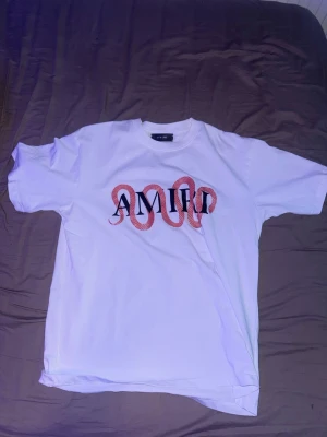 Vit Amiri t-shirt med ormtryck - Säljer en vit t-shirt från Amiri med ett stort svart logotryck på bröstet där en röd orm slingrar sig genom bokstäverna. Klassisk rund hals och korta ärmar. Perfekt statement-plagg för dig som gillar streetwear och coola detaljer.