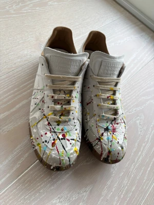 Maison Margiela sneakers med färgstänk - Unika vita sneakers från Maison Margiela med färgstänk i rött, blått, gult och svart. Skorna har klassisk låg siluett, beige snörning och bruna gummisulor. Tillverkade i läder med detaljerad textur på plösen och insida i ljusbrunt läder.