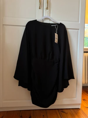 Svart omlottklänning med volang - Svart kortklänning med omlott-design och volangdetaljer. Klänningen har vida, långa ärmar och en draperad effekt framtill och baktill. Tillverkad i ett mjukt, lätt material som ger ett snyggt fall.