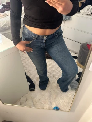 Blå bootcut jeans med låg midja - Säljer ett par blå bootcut jeans med låg  midja och klassisk femficksdesign. Jeansen har en snygg tvätt och raka ben som slutar i en lätt utsvängd passform. Perfekta till en crop top eller hoodie för en chill look.