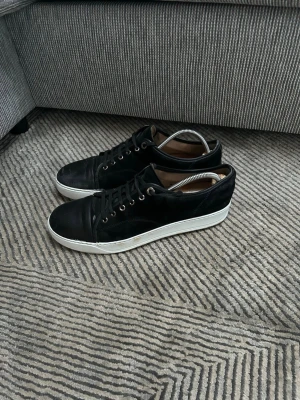 Svarta lanvins  - Stilrena svarta lanvin sneakers med vit sula och svarta skosnören. Skorna har en klassisk låg profil och är tillverkade i läder med metallfärgade öljetter. Perfekta för dig som gillar en clean och enkel look.