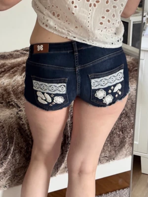Mörkblå jeansshorts med spetsdetaljer - Supersnygga mörkblå jeansshorts med broderade blommor och spets på bakfickorna. Shortsens kanter är fransiga och de har låg midja. Perfekta för sommaren och ger en cool vibe till din outfit.