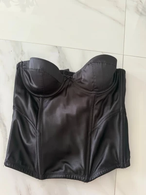 Svart satin korsett med bygel - Snygg svart korsett i glansigt satinmaterial med bygel och vadderade kupor. Modellen har markerade sömmar och ger en riktigt snygg siluett. Perfekt för dig som vill ha en edgy och trendig look.