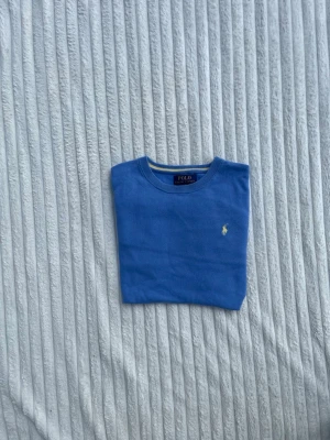 Blå tröja från Polo Ralph Lauren - Snygg blå tröja från Polo Ralph Lauren med rund halsringning och den klassiska broderade loggan i gult på bröstet. Tröjan har långa ärmar och ribbade muddar vid ärmslut och nederkant. Perfekt för en clean och stilren look.