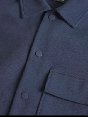 Mörkblå overshirt med tryckknappar - Stilren mörkblå overshirt med klassisk krage och tryckknappar framtill. Jackan har en stor bröstficka med lock och är tillverkad i ett kraftigare tyg som ger en snygg och avslappnad look. Perfekt att bära över en t-shirt för en enkel men trendig stil.