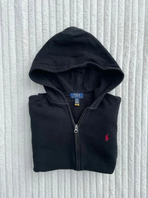 Svart hoodie från Polo Ralph Lauren - Klassisk svart hoodie med dragkedja från Polo Ralph Lauren. Hoodien har huva, kängurufickor och den ikoniska röda loggan broderad på bröstet. Tillverkad i mjukt bomullsmaterial som är skönt att bära. Perfekt för en avslappnad och stilren look.
