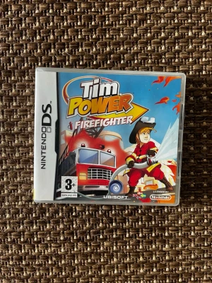 Tim Power Firefighter Nintendo DS - Nintendo DS-spelet Tim Power Firefighter där du spelar som brandman och släcker bränder. Omslaget är färgglatt med en tecknad brandbil och brandman i röd uniform. Spelet är licensierat av Nintendo och rekommenderas från 3 år.