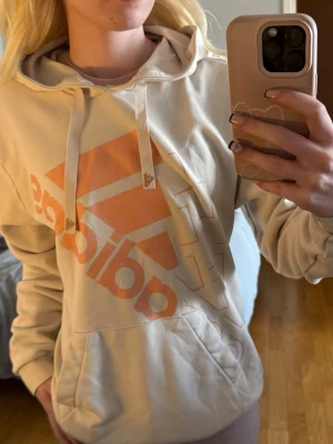 Beige hoodie från Adidas med tryck - Snygg beige hoodie från Adidas med orange/coral logotryck på framsidan. Hoodien har huva med snören och en stor magficka. Tillverkad i mjukt material som känns skönt mot huden. Perfekt för en avslappnad och sportig stil. Finns en liten fläck vid fickan men syns bara på nära håll