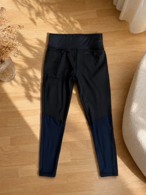 Svarta och marinblå leggings newbody - Snygga leggings från newbody i svart med marinblå detaljer nedtill på benen. Hög midja och praktisk ficka på sidan. Perfekta för träning eller chill. Stretchigt material som sitter skönt och följer kroppen.