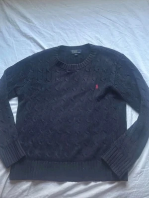 Mörkblå kabelstickad tröja Polo Ralph Lauren - Mörkblå kabelstickad tröja från Polo Ralph Lauren med rund halsringning och röd broderad logga på bröstet. Tröjan har långa ärmar och ribbade muddar vid ärmslut och nederkant. Perfekt för lager på lager och klassisk stil.