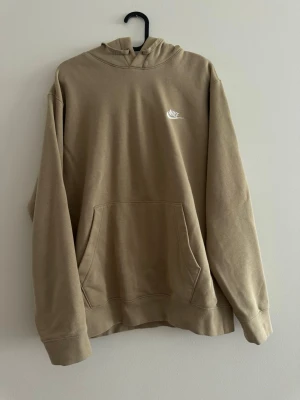 Beige hoodie från Nike med logga - Snygg beige set från Nike med klassisk vit broderad logga på bröstet. Hoodien har stor känguruficka framtill, huva och ribbade muddar vid ärmslut och nederkant. Tillverkad i mjukt bomullsmaterial som känns skönt mot huden. Mjukis med Hoodie