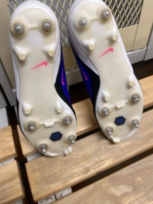 Nike blå och vita fotbollsskor - Säljer ett par Nike fotbollsskor med blå ovandel, vita detaljer och rosa accenter. Skorna har fasta dobbar i metall och plast, snörning och en modern design som ger bra grepp på planen. Perfekta för dig som vill ha stilrena och funktionella skor till matchen.