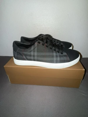 Burberry rutiga svarta sneakers - Snygga svarta sneakers från Burberry med klassiskt rutigt mönster i grått och diskreta röda linjer. Skorna har svarta snören, vit sula och låg profil. Perfekta för dig som gillar stilrena och trendiga skor med exklusiv känsla.