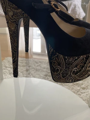 Svarta platåpumps med paisleymönster strl 40 - Unika svarta pumps med hög platå och stilettklack. Skorna har ett elegant paisleymönster i guld och svart på klacken och platån samt en Mary Jane-rem med guldfärgat spänne. Ovandelen är i mockaliknande material för en lyxig känsla.