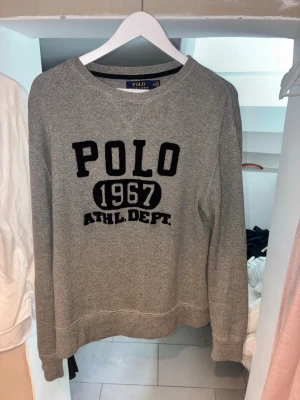 Grå sweatshirt från Polo Ralph Lauren - Cool grå sweatshirt från Polo Ralph Lauren med svart broderad text 'POLO 1967 ATHL. DEPT.' på bröstet. Tröjan har rund halsringning och ribbade muddar. Perfekt för en avslappnad och sportig look.