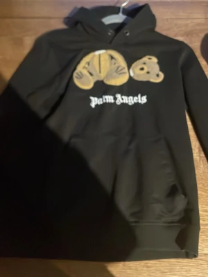Svart Palm Angels hoodie med björn - Svart hoodie från Palm Angels med ikoniskt björnmotiv på bröstet och vit logga under. Klassisk huva och magficka framtill. Tillverkad i mjukt material som passar perfekt för en avslappnad streetwear-look.