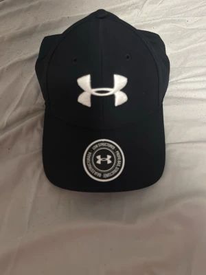 Svart keps från Under Armour - Snygg svart keps från Under Armour med böjd skärm och stort vitt logotryck framtill. Klassisk sexpaneldesign och justerbar passform. Perfekt för en sportig och avslappnad stil.