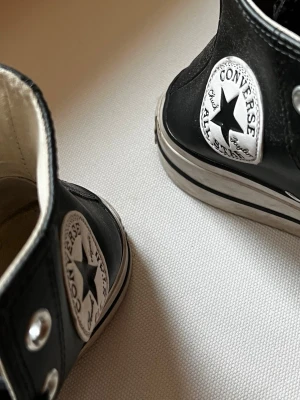 Svarta Converse Chuck Taylor Highs - Klassiska svarta Converse Chuck Taylor All Star med högt skaft, vita gummisulor och tåhättor. Skorna har svarta skosnören, metallöljetter och det ikoniska Converse-märket på sidan. Ovandelen är i canvas och insidan är vit.