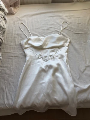 Vit satin klänning med smala axelband - Snygg vit kort klänning i satin med smala axelband. Klänningen har en enkel och elegant design, perfekt nu på sommaren. Den är aldrig använd då den är lite för stor på mig. 