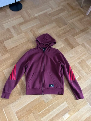 Vinröd hoodie från Adidas med dragkedja - Säljer en vinröd hoodie från Adidas med dragkedja framtill och huva. Tröjan har de klassiska tre röda ränderna längs båda ärmarna och en liten Adidas-logga på magen. Tillverkad i mjukt bomullsmaterial och har två fickor framtill. Har matchande byxor på min profil:)