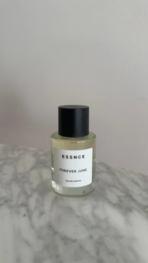 ESSNCE Forever June parfym - Minimalistisk parfymflaska från ESSNCE med doften Forever June. Flaskan är cylindrisk i genomskinligt glas med svart rund kork och vit etikett med svart text. Perfekt för dig som gillar stilrena och moderna detaljer på parfymhyllan. Helt ny aldrig använd 