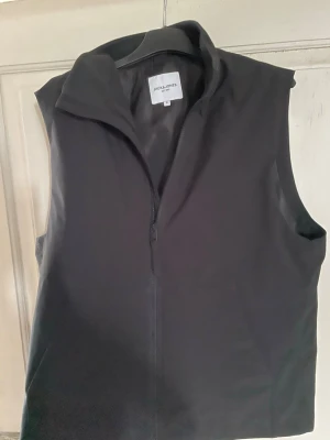 Svart väst från Jack & Jones - Stilren svart väst från Jack & Jones med hög krage och dragkedja framtill. Perfekt att slänga över en hoodie eller tröja för en clean look. Västen är enkel och passar till det mesta i garderoben.