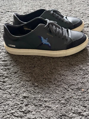 Axel Arigato Sneakers - Snygga svarta sneakers från Axel Arigato med broderad blå fågel på sidan och vit sula. Tillverkade i läder med mockadetaljer vid tån. Perfekta för dig som gillar stilrena och trendiga skor.