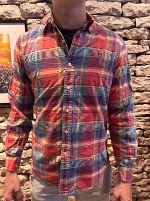 Rutig skjorta från Polo Ralph Lauren - Säljer en färgglad rutig skjorta från Polo Ralph Lauren med klassisk button-down krage och lång ärm. Skjortan har en mix av rött, blått, grönt och gult i ett rutmönster samt den ikoniska broderade loggan på bröstet. Tillverkad i mjuk bomull.