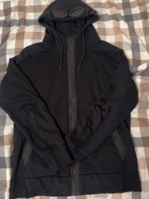 Svart hoodie med goggles från C.P. Company - Svart hoodie från C.P. Company med hel dragkedja och två fickor framtill. Huvan har inbyggda goggles och dragsko. Tillverkad i mjukt material, perfekt för en streetwear-look.