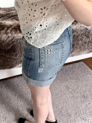 Blå jeanskjol med nitar - Coola blå jeansskorts med uppvikt kant och slitningar. Skortsen har klassiska fickor och silverfärgade nitar på bakfickan för en edgy look. Perfekt för dig som vill ha både komfort och stil. OBS: litet svagt märke, se sits bild