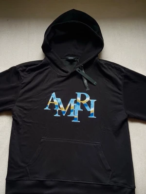 Svart AMIRI hoodie med blå logga - Snygg svart hoodie från AMIRI med stor blå och gul logga på bröstet. Pris kan diskuteras vid snabb affär 