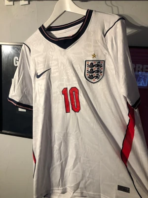 England fotbollströja Bellingham #10 - Säljer en vit England fotbollströja från Nike med Bellingham och nummer 10 i rött på ryggen. Tröjan har broderad landslagssköld, gul stjärna ovanför och marinblå detaljer vid krage och ärmslut. Tillverkad i lätt och ventilerande material, perfekt för match eller träning.
