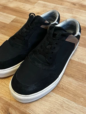 Svarta burberry sneakers med mönster - Snygga svarta burberry sneakers med vit sula och rutiga detaljer i svart, vitt och brunt på hälen. Ovandelen är i mocka och textil, med svarta skosnören och låg profil. Perfekta för dig som gillar stilrena men ändå unika skor.