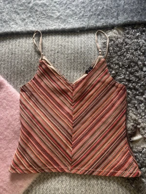 Randig linnetopp med smala axelband - Säljer en färgglad randig linnetopp med smala axelband och v-ringad hals. Toppen har ett mönster i rosa, orange, beige och brunt med tunna ränder i olika färger. Perfekt för varma dagar och enkel att styla till jeans eller kjol.