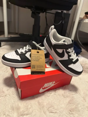 Svarta och vita Nike Dunk sneakers - Snygga Nike Dunk sneakers i svart och vitt med klassisk låg siluett. Skorna har vit tåbox, svarta detaljer och Nike swoosh på sidan. Tillverkade med minst 20% återvunnet material. Perfekta för dig som gillar streetwear och vill ha en clean look.