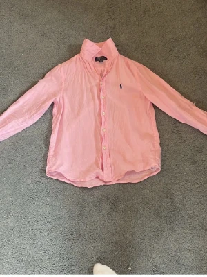 Rosa barn skjorta från Polo Ralph Lauren - Säljer en klassisk rosa skjorta från Polo Ralph Lauren med lång ärm och knappar framtill. Skjortan har den ikoniska broderade loggan på bröstet och är tillverkad i mjuk bomull. Perfekt till jeans eller chinos för en clean look.