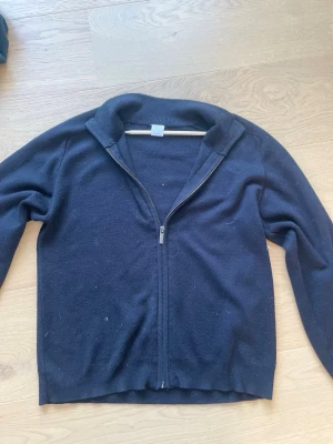 Svart ziptröja från Lindex Kids - En enkel svart ziptröja från Lindex Kids med hel dragkedja framtill och ribbade muddar. Perfekt att slänga på sig över en t-shirt. Tröjan har en klassisk passform och är gjord i mjukt material.