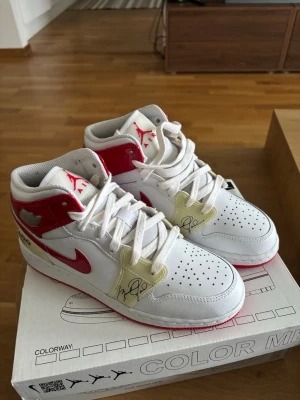 Air Jordan 1 Mid Chicago White/Red - Säljer ett par Air Jordan 1 Mid sneakers i klassisk Chicago-färg. Skorna är vita med röda detaljer och beige paneler med tryck. Ikonisk röd swoosh och Jordan-logga på plösen. Perforerad tå och vita skosnören. Perfekta för dig som gillar streetwear och sneakerskultur.