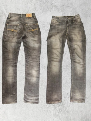 Vintage Nudie Jeans Thinn Finn - Sällsynta vintage Nudie Jeans i modellen Thinn Finn (äldre stavning med dubbelt N) – troligtvis från tidigt 2000-tal. 100% bomull utan elastan, vilket är typiskt för Nudies äldsta produktion och ovanligt idag.  Colorwayen är en djup brun/grå ton med kraftiga naturliga fades, whiskering vid låren och ljusare partier – klassisk inbyggd karaktär som inte går att replikera.  Mått:  	•	Midja: 37 cm 	•	Längd: 109 cm 	•	Benöppning: 18 cm (±1 cm)  Passar ca W29/L32.