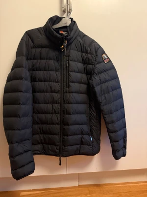 Svart dunjacka från Parajumpers - Snygg svart dunjacka från Parajumpers med quiltad design och hög krage. Jackan har dragkedja framtill, en bröstficka med dragkedja och logotyp på ärmen. Perfekt för kalla vinterdagar och riktigt stilren look. Som ny skick inga flaws 