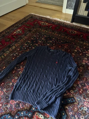 Mörkblå kabelstickad tröja Polo Ralph Lauren - Kabelstickad mörkblå tröja från Polo Ralph Lauren med rund halsringning och det klassiska röda broderade logot på bröstet. Långa ärmar och ribbade muddar vid ärmslut och nederkant. Perfekt för lager på lager och snygg till jeans.