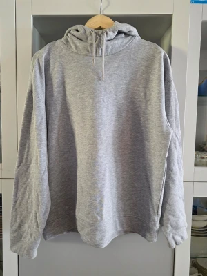 Grå basic hoodie med huva - En stilren grå hoodie med huva och dragsko. Tillverkad i mjukt och bekvämt material, perfekt för en avslappnad look. Hoodien har långa ärmar och ribbade muddar vid ärmslut och nederkant. Passar dig som gillar enkel och klassisk streetstyle.