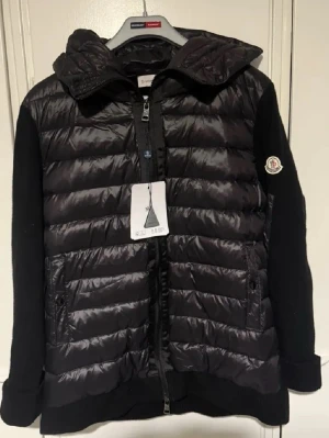 Svart pufferjacka från Moncler - Snygg svart pufferjacka från Moncler med quiltad front och huva i glansigt material. Ärmar och rygg är i mjuk stickad kvalitet. Jackan har dragkedja framtill, två fickor och Moncler-logga på ärmen. Perfekt för kalla vinterdagar och riktigt trendig look.