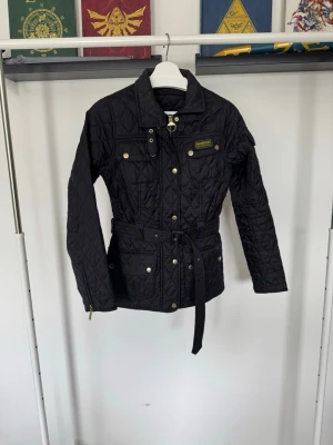 Svart quiltad jacka från Barbour - Säljer en svart quiltad jacka från Barbour med bälte i midjan, guldfärgade knappar och dragkedja. 