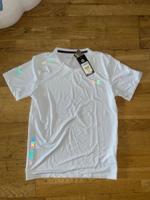 Vit Under Armour t-shirt med reflex logga - Snygg vit t-shirt från Under Armour med holografiska loggor och textdetaljer på bröstet, ärmen och vid nederkanten. Klassisk rund hals och korta ärmar. Perfekt för dig som gillar sportig och clean stil. Det står storlek M men det passar både M och S.