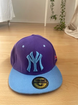 Lila och blå YMCMB keps - Snygg keps i lila med ljusblå skärm och broderad NY-logga framtill. Kepsen har även små ljusblå detaljer och en röd broderad fågel på sidan. Perfekt accessoar för att lyfta din streetwear-look.