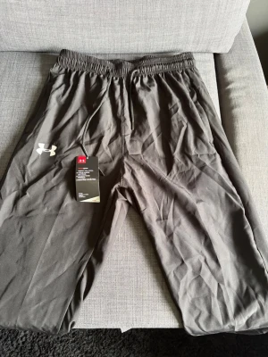 Svarta polyester byxor från Under Armour - Snygga svarta polyester byxorfrån Under Armour med dragsko i midjan och fickor med dragkedja. Tillverkade i ett mjukt och lätt material som känns skönt mot huden. Perfekta för chill eller träning. Klassisk passform med resår i midjan.