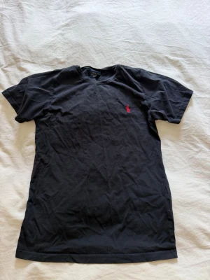 Svart t-shirt från Polo Ralph Lauren - Klassisk svart t-shirt från Polo Ralph Lauren med röd broderad logga på bröstet. T-shirten har rund halsringning och korta ärmar. Tillverkad i mjuk bomull för en bekväm känsla och enkel stil.