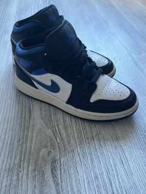 Air Jordan 1 Mid svart/blå/vit - Säljer ett par Air Jordan 1 Mid sneakers i svart, blått och vitt. Klassisk siluett med perforerad tåbox, svarta skosnören och blå detaljer på swoosh och logga. Ovandelen är i läder och yttersulan är vit med svart kant. Perfekta för dig som gillar streetwear och ikoniska sneakers.